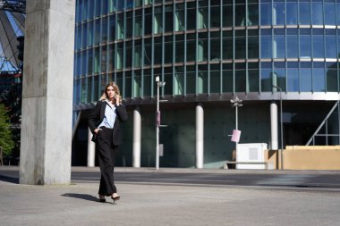Takım elbiseli genç bir kadın sokakta yürüyor ve cep telefonuyla konuşuyor. İş kadını işe giderken telefonla konuşuyor..