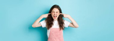 Sinirli genç esmer kadın çığlık atıyor ve kulaklarını kapatıyor, sinir bozucu sesi engelliyor, birini görmezden geliyor, korkunç sesler duyuyor, mavi arka planda duruyor..