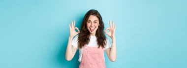 Kıvırcık saçlı heyecanlı esmer kızın portresi, iyi işaretler gösteriyor, mükemmel seçimi övüyor, iyi bir şey öneriyor, tamam işareti veriyor, mavi arka planda duruyor..