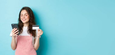 Genç, modern bir kız kredi kartıyla ödeme yapıyor, akıllı telefondan bilgi yazıyor, mutlu bir yüz ifadesiyle ekrana bakıyor, uygulamadan alışveriş yapıyor, mavi arka planda duruyor..