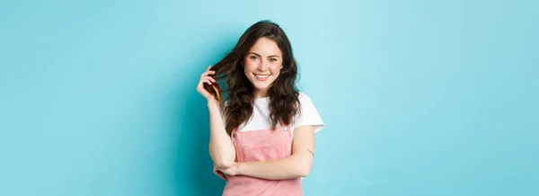 stock image Beautiful teenage girl waiting for summer, playing with curly hair and smiling white teeth, having cute blush and glam make up on face, standing against blue background.