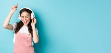 Milenyum kızı bahar kıyafetiyle dans ediyor, kulaklıkla müzik dinliyor ve sesten zevk alıyor, mavi arka planda duruyor..