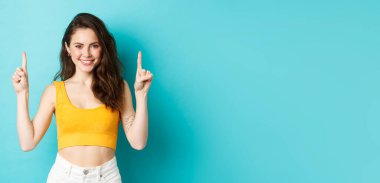 Yaz yaşam tarzı konsepti. Kendine güvenen genç, şık bir kadın, sarı bir bluz giyiyor, fotokopi alanını işaret ediyor, mavi arka planda duruyor..