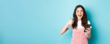 Heyecanlı şirin kızın portresi evet de, gülümse ve onaylamak için başını salla, parmaklarını gösterirken akıllı telefonu tut, ve güzel online teklifi öv, mavi arka planda dur..