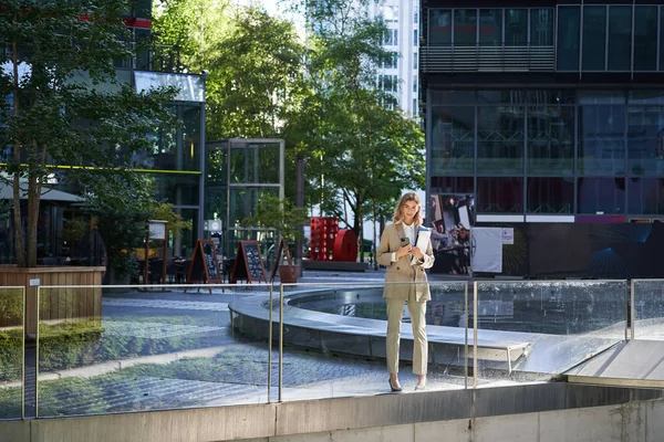 Corporate woman in suit, standing with confidence in beige suit with working documents outdoors, posing in city center.