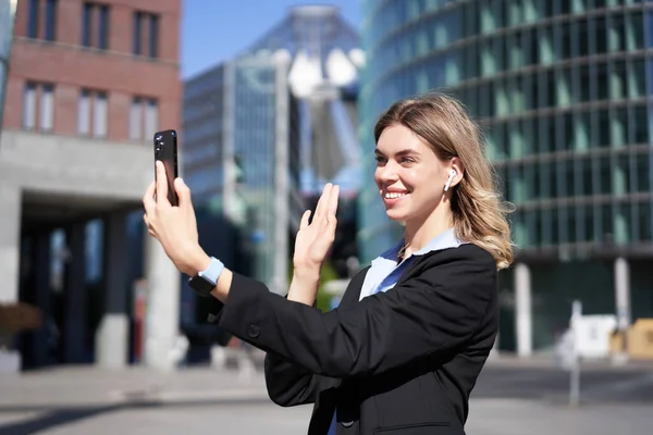 Portrait of businesswoman wave her hand at mobile phone camera, waves hand during video chat, stands in suit in city center outdoors.