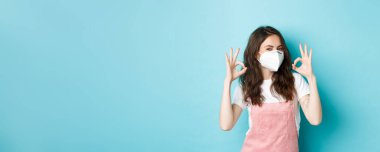 Covid, sağlık ve salgın hastalık konsepti. - Çok iyi. Tıbbi solunum cihazı takan genç destekçi bir kadın onaylandığına dair belirtiler gösteriyor, halk içinde maske takıyor, mavi arka plan.