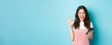 Heyecanlı ve memnun genç bir kadının görüntüsü akıllı telefon uygulaması kullandıktan sonra evet diyor, cep telefonuyla online alışveriş yapıyor ve tamam işareti gösteriyor, mavi arka planda duruyor..