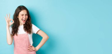 Neşeli cazibeli bir kadının göz kırpması ve iyi bir şeyi tavsiye etmek ve onaylamak için tamam işareti göstermesi, evet demek, iyi seçimi övmek, mavi arka plan..