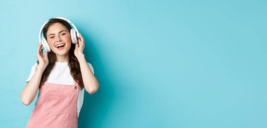 Modern kız kablosuz kulaklıkla müzik dinliyor, kulaklıklara dokunuyor, gülümsüyor, şarkı söylüyor, mavi arka planda duruyor..