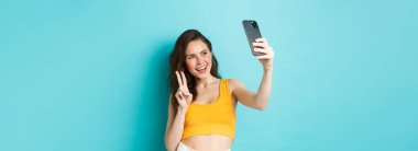 Teknoloji ve yaşam tarzı konsepti. Şık genç bir kadın yaz kıyafetleriyle selfie çekiyor, akıllı telefon kamerasında V işareti gösteriyor, mavi arka planda duruyor..