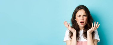 Kafası karışmış, kaşlarını çatmış, suratını buruşturmuş, yüzünü buruşturmuş, anlamayan, mavi arka planda dikilen esmer kızın yakın portresi..