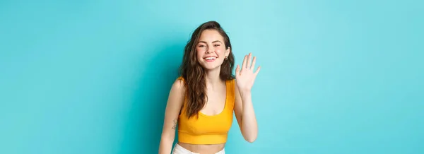 Summer holidays and emotions concept. Beautiful friendly woman with perfect body, waving hand and saying hi, greeting you, standing over blue background.