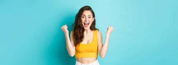 Summer holidays and emotions concept. Excited young woman winning and screaming from joy, achieve goal, celebrating victory, making fist pumps, standing over blue background.