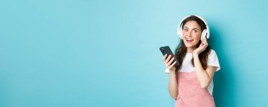 Kulaklık takan, akıllı telefondan müzik çalan, telefon tutan, şarkı dinleyen, mavi arka planda duran genç bir kadın görüntüsü..