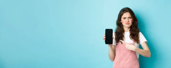 Skeptical and upset young woman frowning, sulking disappointed while pointing at empty smartphone screen with bad online offer or app, standing against blue background.
