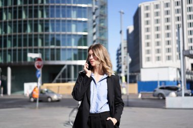 Takım elbiseli bir iş kadını portresi, sokakta cep telefonuyla dikiliyor, telefonla konuşurken gülümsüyor..