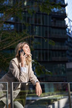 Bej takım elbiseli kendinden emin genç bir kadın, cep telefonuyla konuşan ve güneşli bir günde gülümseyen bir şirket hanımı,.