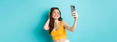 Güzel modern kız tatilde selfie çekiyor, yaz kıyafetleriyle poz veriyor ve akıllı telefon kamerasına bakıyor, mavi arka planda duruyor..
