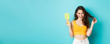 Mutlu, çekici bir kadın saç teli olmadan fırçasını gösteriyor ve uzun, sağlıklı bir saç stiline dokunuyor. Mutlu bir yüzü var. Şampuan sonuçlarından memnun, mavi arka plan..