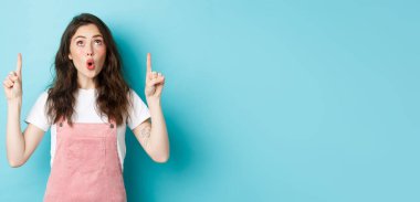 Vay canına, çok havalı. Siyah kıvırcık saç stili ve cazibeli etkileyici genç kız, şaşkın ve heyecanlı görün, reklamlara işaret et ve dik dik bak, mavi arka plan.