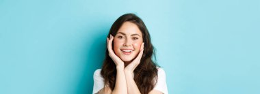 Güzel bir kadının parıldayan tenine dokunduğu, makyaj yaptığı, dişleriyle gülümsediği, mavi arka planda dikildiği yakın plan..