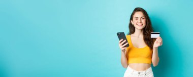 İnternetten alışveriş. Güzel bir kadın yaz tatiline hazırlanıyor, biletleri kredi kartı ve akıllı telefon uygulaması ile ayırtıyor, mavi arka planda duruyor..