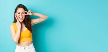 Heyecanlı kız sevinçten bağırıyor, yaz tatiline hazır, V işaretini gösteriyor ve gülümsüyor, mavi arka planda mutlu duruyor..