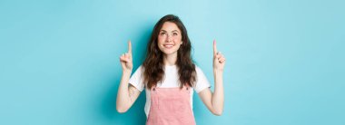 Çekici makyajlı genç bir kız, pembe yaz tulumları giyiyor, parmaklarını kaldırıp gülümsüyor, en tepedeki tanıtım teklifine bakıyor, mavi arka plan..