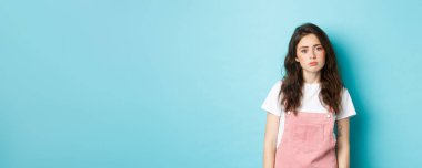 Üzgün ve yorgun genç kadın görüntüsü yalnız ve üzgün, iç çekiyor, kameraya bakıyor, mavi arka planda duruyor..