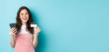 İnternetten alışveriş yapan, plastik kredi kartı gösteren, akıllı telefon tutan, mavi arka planda duran şık genç bir kadın..