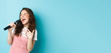 Kaygısız güzel kız şarkı söylüyor, mikrofonda tutkuyla şarkı söylüyor, karaoke yapıyor, mavi arka planda duruyor..