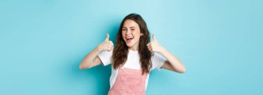 Kıvırcık saçlı, gülümseyen genç bir kadının portresi, onaylanmış parmaklarını gösteriyor, evet diyor, tavsiye veriyor, övgüler yağdırıyor, mavi arka planda duruyor..