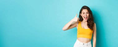 Yaz tatili ve duygu konsepti. Neşeli genç bir kadın evet diyor, iyi seçimi övüyor, iyi iş çıkarıyor, başparmaklarını kaldırıyor ve heyecanlı görünüyor, mavi arka planda duruyor..