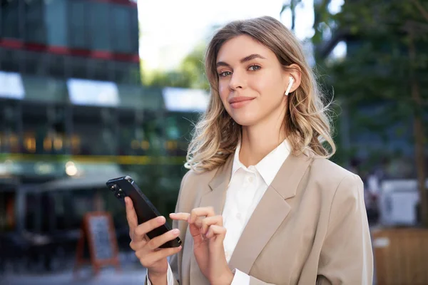 Smiling beautiful corporate woman in beige suit, standing on street in city with wireless headphones, holding smartphone.