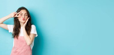 Romantik ve güzel genç bir kadın havada öpüşüyor, dudaklarını büzüyor ve mavi arka planda dikilirken göze yakın V işareti hareketi gösteriyor..