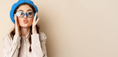 Beautiful stylish woman in sunglasses, blowing air kiss on hands near lips, gazing softly and flirty at camera, posing against beige background.