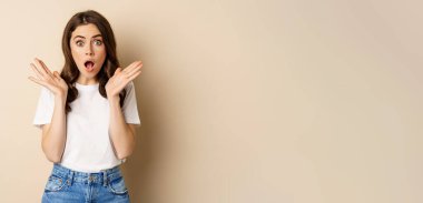 Portrait of stylish young woman gasping, looking surprised and amazed, impressed by smth, standing in t-shirt and jeans over beige background.