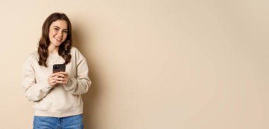 People and cellular technology. Beautiful stylish woman using mobile phone, smartphone app, smiling and looking at screen, standing over beige background.