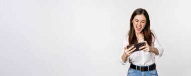 Happy brunette woman playing mobile video game, smiling and looking at screen excited, standing over white background.