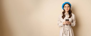 Image of young stylish french woman in trench coat, standing with smartphone, using mobile phone app and smiling, posing against beige background.
