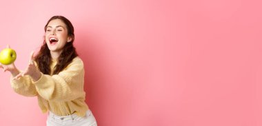 Mutlu tatlı kız bir elma yakalar ve güler, sağlıklı öğle yemeği zamanı meyvelerle dolu, pembe arka planda durur.