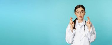 Hopeful woman doctor, lab worker cross fingers and smiling, praying, making wish, anticipating, standing over torquoise background.