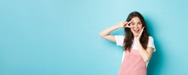 Heyecanlı çekici bir kadının portresi, göze yakın V işareti gösteriyor, Kawaii 'yi mutlu ediyor ve kameraya gülümsüyor, mavi arka plana karşı neşeli duruyor..