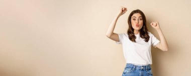 Enthusiastic adn surprised woman winning, dancing and celebrating, standing in tshirt over beige background.
