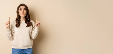 Amazed girl checking out banner announcement, reading promo text upwards, pointing fingers up, standing over beige background.