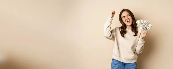 Enthusiastic modern woman winning money, got cash, celebrating and shouting of joy, standing against beige background.