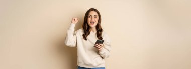 Happy brunette girl holding smartphone and cheering, winning, celebrating win on mobile phone app, standing over beige background.