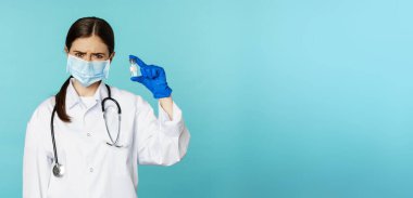 Üzgün ve hayal kırıklığına uğramış maskeli kadın doktor, aşıyı üzgün bir suratla gösteriyor, mavi arka planda üniformalı..