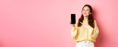 Çekici genç bir kadının portresi telefonda tanıtımını gösteriyor, akıllı telefonu işaret ediyor ve gülümsüyor, pembe arka planda duruyor..